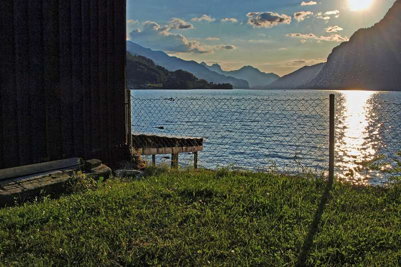 Blick über den Walensee im hellen Sonnenlicht. Im Vordergrund befindet sich eine grüne Wiese mit einem Maschendrahtzaun, links ragt die dunkle Wand einer Holzhütte ins Bild. Im Hintergrund sind Gebirgsketten und Himmel mit wenig Wolken zu sehen.