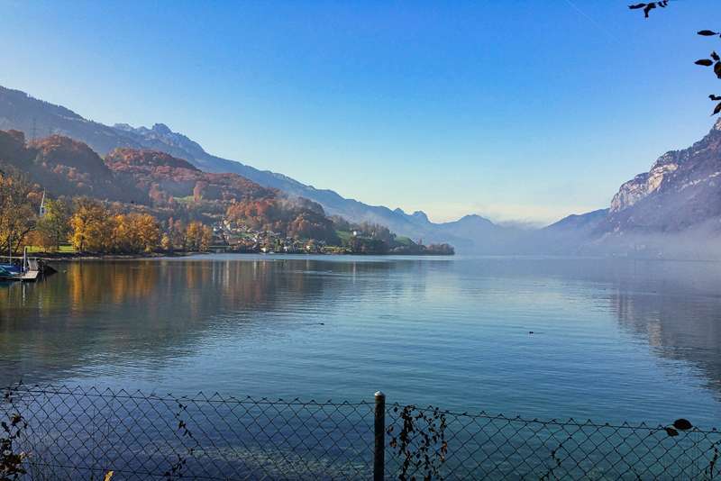Herbstliche Idylle am See: Während sich die bunten Wälder und Hänge am linken Ufer im spiegelglatten Wasser reflektieren, zieht auf der rechten Seite sanfter Nebel an den steilen Felswänden vorbei.