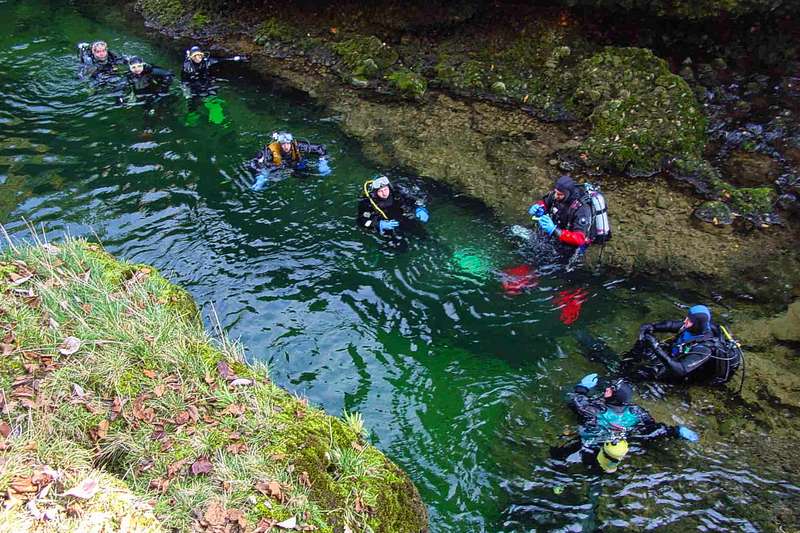 Eine Gruppe von Sporttauchern bereitet sich in einem idyllischen, grün schimmernden Flussbett auf den Tauchgang vor.